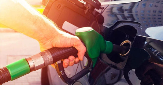 A close-up shot of a hand holding a green fuel nozzle and inserting it into the gas tank of a black car. The scene is bright with sunlight streaming in from the left.
