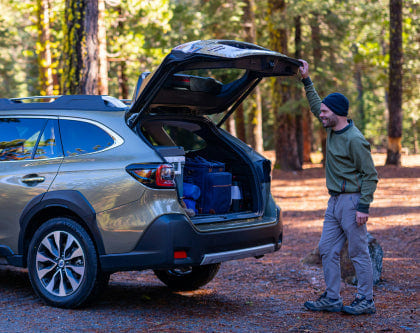 A man in green clothes, grey pants and a hat is opening the trunk of a car to put his belongings in.